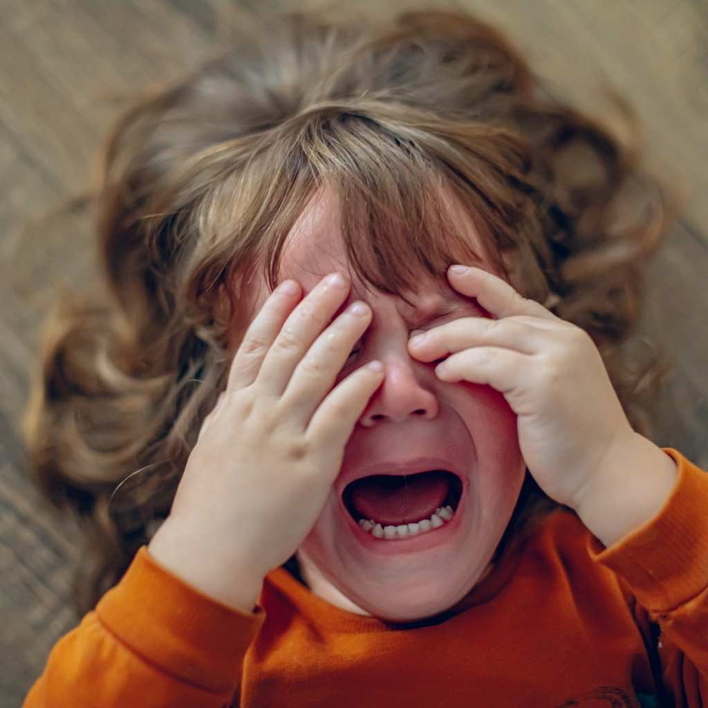 A young child lying on the floor with hands covering their face while crying.