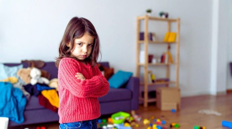 A young girl with crossed arms and an angry expression stands in a cluttered living room, illustrating the challenges of autism and anger, including frustration and sensory overload.
