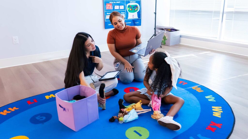 The Essential Role Of A Bcba In Autism Therapy: 3 Important Facts Bcba observing an aba therapy session while a therapist interacts with a young girl playing with dolls on a colorful alphabet rug in a clinic room