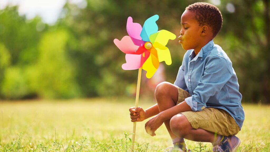 Young child crouching in the grass and blowing on a colorful pinwheel, representing childhood development and autism diagnosis.