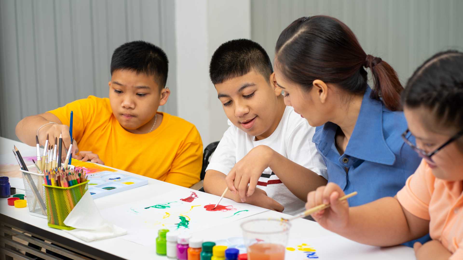 Autism support groups in Texas: Three children are sitting around a table painting; a woman is instructing the middle child.