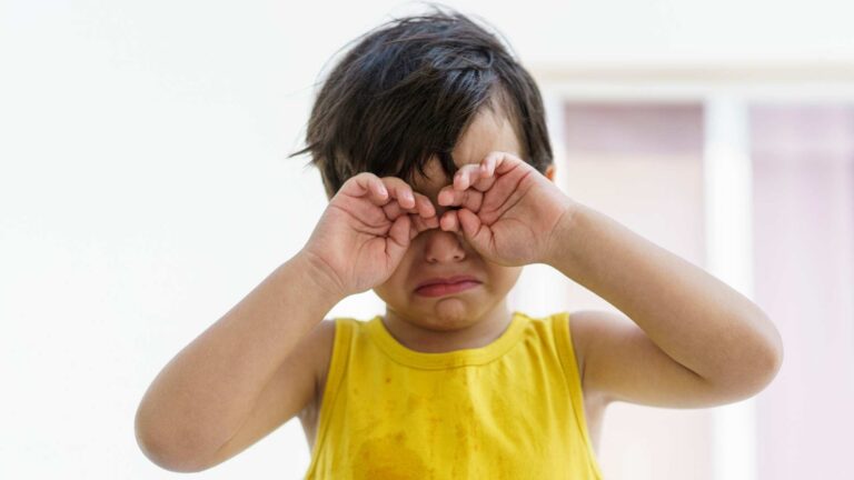 A young child wearing a yellow shirt holding their hands over their eyes as if upset.