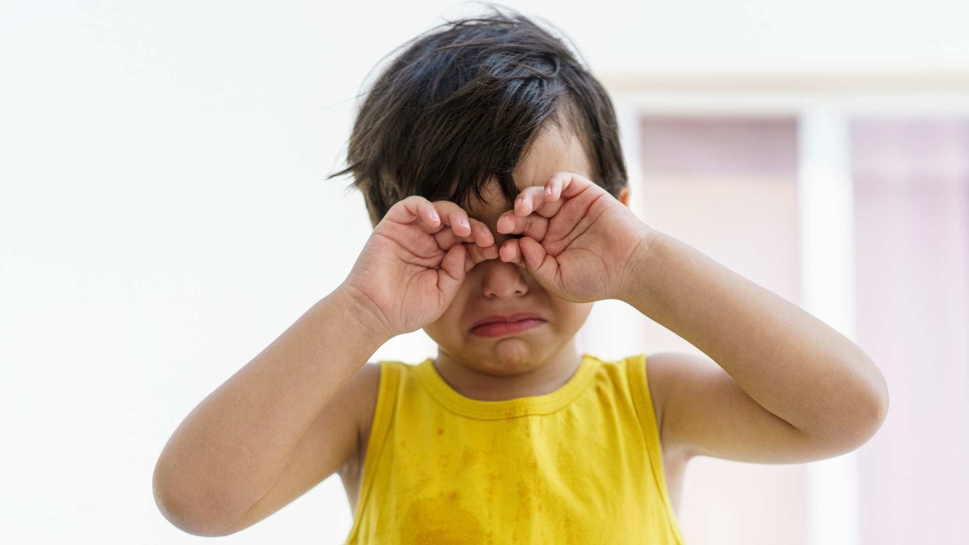 A young child wearing a yellow shirt holding their hands over their eyes as if upset.