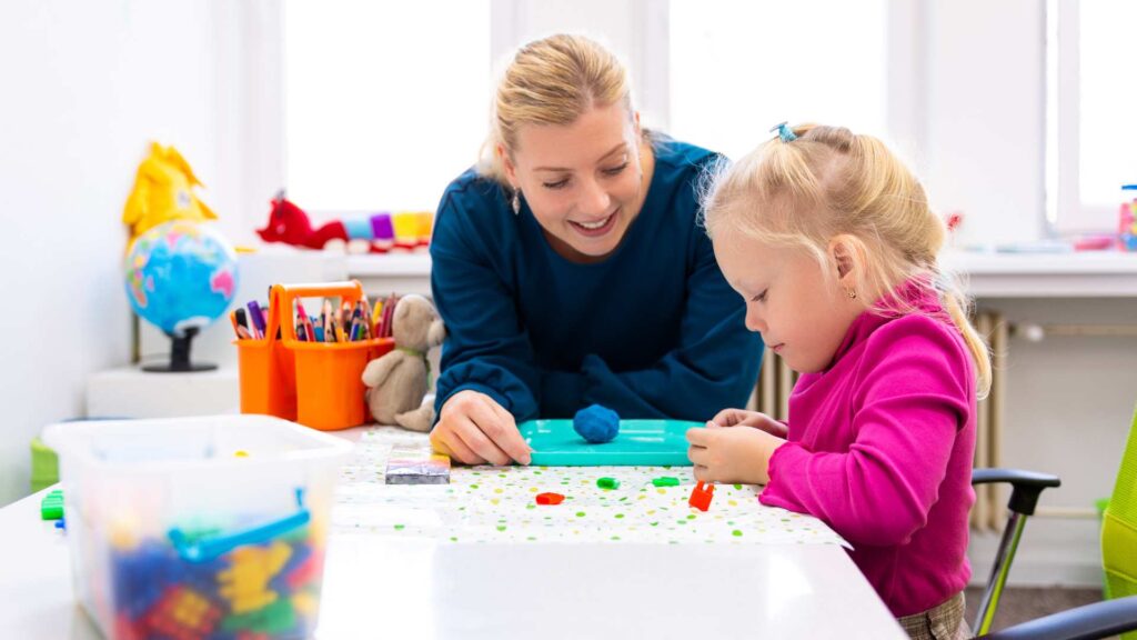 Therapist guiding a young child through a colorful tabletop activity during an ABA therapy session in a bright classroom