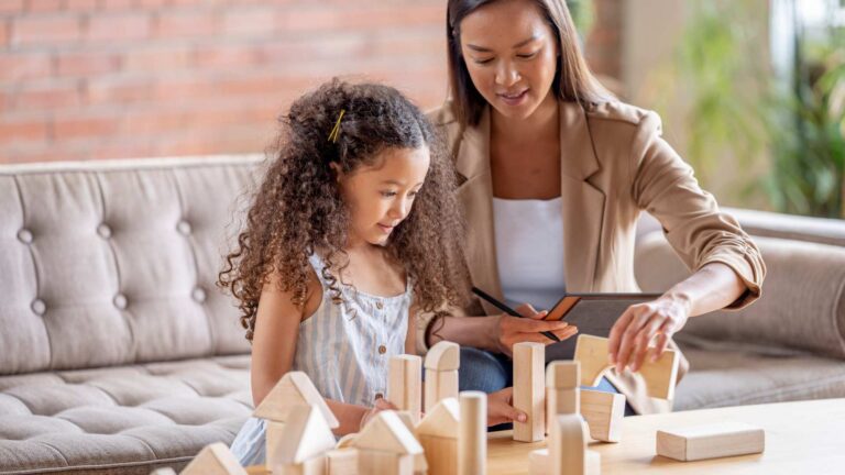 Therapist sitting beside a child on a sofa table, using a tablet and blocks to practice listening, cooperation, and praise as soft skills in ABA Therapy