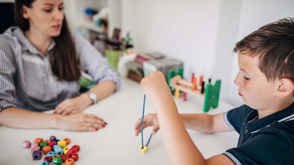 Therapist supporting a young boy in a structured aba activity, threading beads to build focus and coordination