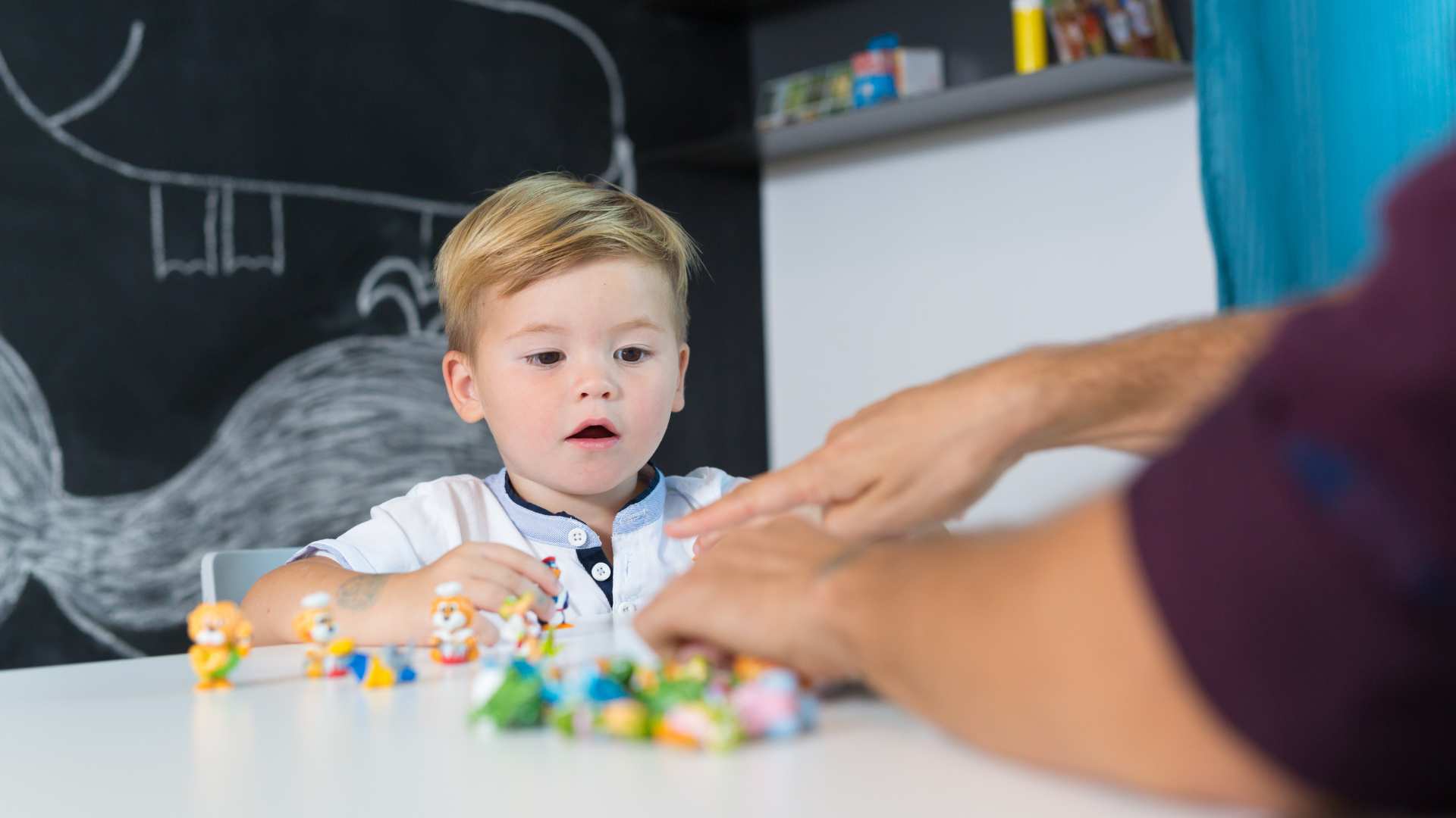 Toddler boy focuses on small toy figures on a table while a therapist guides him during an ABA therapy for toddlers session in a clinic room