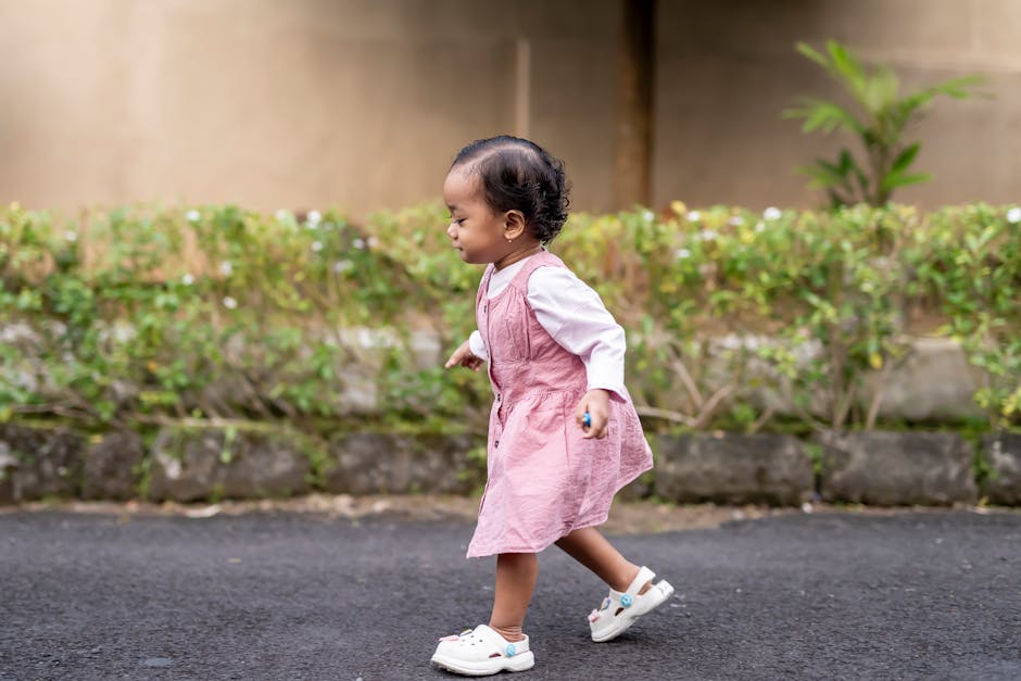 Cute toddler in a pink dress walking on a pathway with green foliage background, showing early steps.