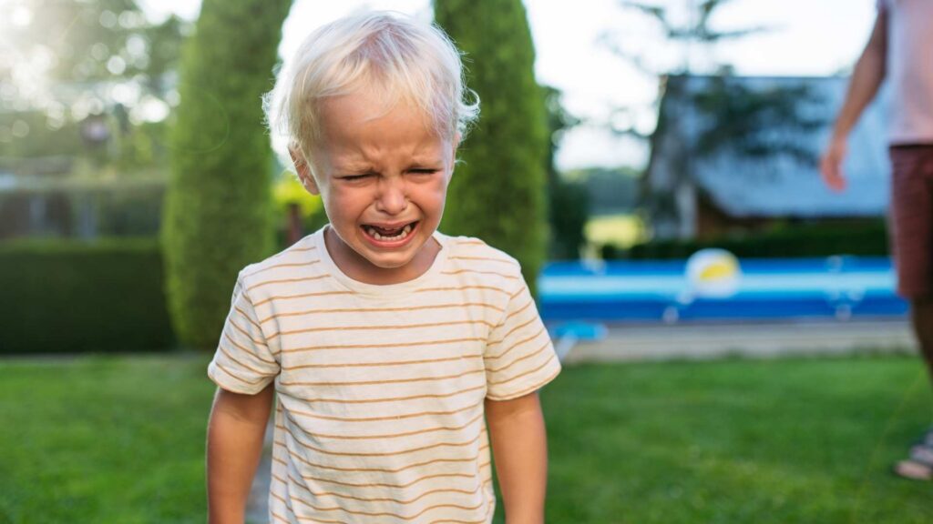 A young child outdoors crying on a sunny day, standing on green grass near a pool.